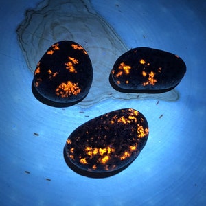 May include: Three smooth, dark gray stones with bright orange flecks, illuminated under a UV light. The stones are arranged in a triangular pattern on a blue surface.