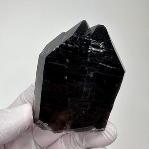 Może przedstawiać: Ciemny, nieprzezroczysty, czarny okaz kryształu o fasetowanej, geometrycznej strukturze. Minerał posiada ostre, kanciaste krawędzie oraz gładką, odblaskową powierzchnię z widocznymi wewnętrznymi pęknięciami i naturalnymi wzorami.