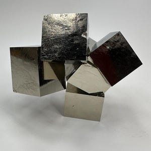 Op de afbeelding: Een cluster van metalen pyrietkubussen, met een reflecterend, zilvergrijs oppervlak. De kubussen zijn gerangschikt in een dynamische, overlappende compositie, met zichtbare kristallijne structuren. De algehele esthetiek is modern en geologisch.