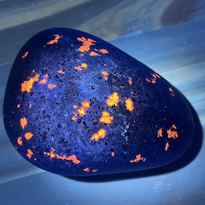 Puede incluir: Una roca ovalada lisa de color azul oscuro con motas de color naranja brillante. La roca está iluminada con luz UV, lo que hace que las motas naranjas brillen.