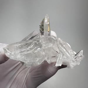 Pode incluir: Um aglomerado de cristais de quartzo claros e translúcidos, apresentando múltiplas terminações afiadas e pontiagudas, além de superfícies facetadas irregulares. O espécime mineral exibe vários graus de transparência e fraturas internas.