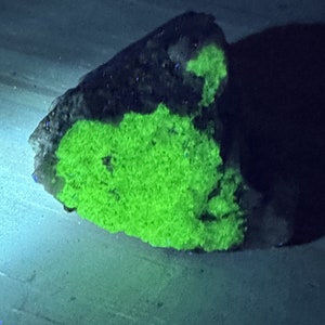 Op de afbeelding: Een donkergrijze rots met een felgroene mineraalafzetting erop. Het mineraal gloeit onder een zwart licht.