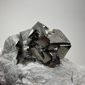 Könnte beinhalten: Ein großer, metallischer, grauer Pyritkristall-Cluster auf einem grauen Stein. Der Kristall hat eine kubische Form und ist sehr reflektierend.