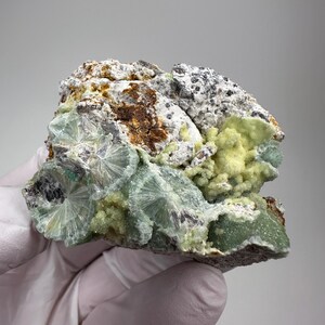 May include: A raw mineral specimen featuring pale green, radiating crystal clusters alongside patches of bright yellow, granular material. The rock matrix displays a mix of white, grey, and brown earthy textures with rough, uneven surfaces throughout.