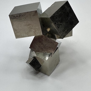 Op de afbeelding: Een cluster van natuurlijke pyrietkristallen met meerdere onderling verbonden metalen kubussen met een reflecterend, messingkleurig gouden oppervlak. Het mineraal vertoont scherpe geometrische randen en gladde vlakken in een complexe, driedimensionale formatie.