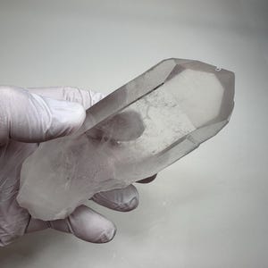 May include: A large, clear quartz crystal with a pointed tip. The crystal is held in a gloved hand, showcasing its natural, raw form. The quartz has a translucent appearance with subtle internal patterns, reflecting light.