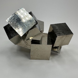Könnte beinhalten: Ein Cluster aus metallischen Pyritwürfeln, angeordnet in einer scheinbar zufälligen, aber strukturierten Formation. Die Würfel haben eine reflektierende, silbergraue Oberfläche, die das Licht einfängt und Glanzlichter und Schatten erzeugt. Der Gesamteffekt ist eine moderne, geometrische Skulptur.