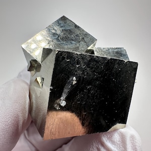 Könnte beinhalten: Ein metallisches Pyrit-Mineralspecimen mit ausgeprägten kubischen Kristallformationen und einer reflektierenden, messinggoldenen Oberfläche. Die Gruppe zeigt scharfe geometrische Kanten und glatte, spiegelnde Flächen mit kleinen Einschlüssen.