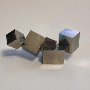 Op de afbeelding: Een cluster van metalen pyrietkristallen met verschillende onderling verbonden, natuurlijk gevormde kubussen met een reflecterend, messinggoud oppervlak. Het mineraal vertoont scherpe, geometrische randen en gladde, platte vlakken.