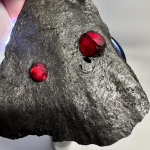Op de afbeelding: Een driehoekig stuk donkergrijs, getextureerd gesteente met twee ingebedde, cirkelvormige, felrode edelstenen. Het oppervlak van de steen is ruw en ongelijkmatig, wat contrasteert met het gladde, gepolijste uiterlijk van de twee rode kristallen.