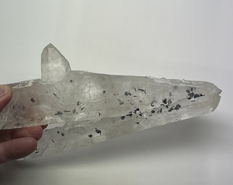 May include: A large, clear quartz crystal with black inclusions. The crystal is shaped like a point and has a rough, natural surface.
