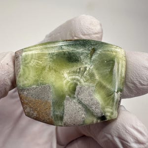 May include: A polished, rectangular gemstone with a light green and gray pattern. The stone has a unique, swirling design in shades of green, with areas of gray and brown. The edges are slightly rounded, and the stone appears to be smooth and glossy.