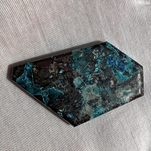 May include: A dark blue and green gemstone with a rough, irregular shape. The gemstone has a mottled appearance with a mix of dark and light blue and green colors.