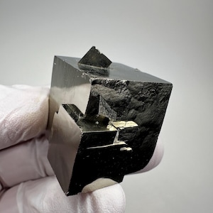 Op de afbeelding: Een donkere, metalen pyrietkubus met een glanzend, reflecterend oppervlak. Het mineraalmonster heeft een geometrische vorm met een kleine piramidale formatie aan de bovenkant. De pyriet wordt vastgehouden door een gehandschoende hand tegen een neutrale achtergrond.