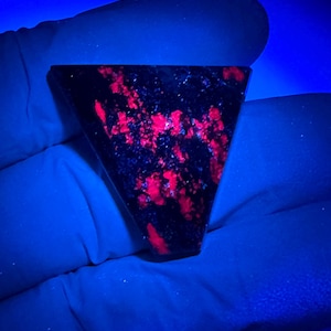 May include: A triangular-shaped stone cabochon featuring a dark, near-black base color with vibrant, glowing red inclusions that fluoresce under ultraviolet light. The surface is smooth and polished, displaying a distinct geometric triangular form.