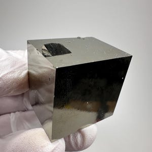 Puede incluir: Un cubo de pirita pulida, también conocida como oro de los tontos, con brillo metálico. El cubo es sostenido por una mano enguantada sobre un fondo blanco. La pirita tiene una superficie reflectante y una forma geométrica.