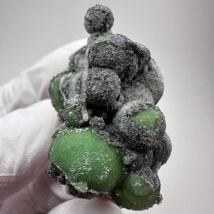 May include: A botryoidal mineral specimen featuring clusters of smooth, vibrant green, rounded formations partially coated in a textured, dark grey to black crystalline crust. The structure displays a grape-like appearance with varying sizes of spheres.