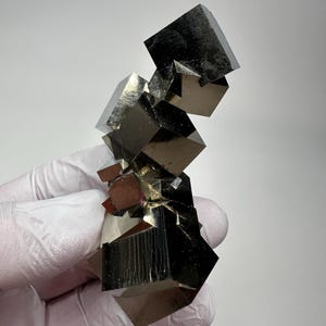 以下が含まれることがあります： 鋭い立方体の結晶が連なった金属質の黄鉄鉱のクラスター。反射する真鍮のような金色の表面を持ち、幾何学的なエッジと滑らかで平らな面が光を捉えます。個々の立方体の間には深い影があり、複雑で多面的な構造を形成している鉱物標本です。