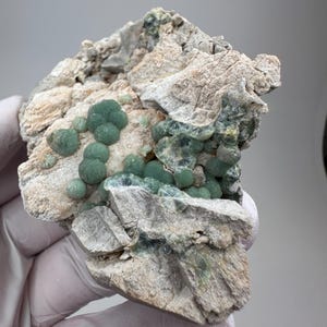 May include: A mineral specimen featuring clusters of small, spherical, forest green crystals embedded within a rough, light beige, and textured rocky matrix. The green formations exhibit a radial, fibrous structure with some translucent areas.