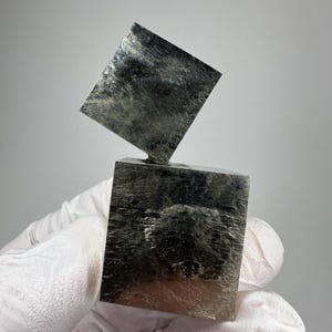 May include: Two metallic, brass-coloured pyrite cubes stacked at an angle. The mineral specimens feature a reflective, crystalline surface with natural striations and sharp, geometric edges, showcasing a distinct cubic structure and metallic lustre.