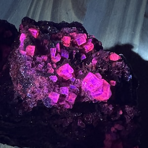 May include: A close-up of a dark purple rock with bright pink crystals. The crystals are clustered together and have a shiny, reflective surface.