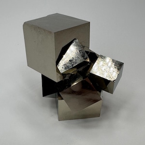 May include: A cluster of metallic pyrite crystals featuring multiple interconnected cubic formations with smooth, reflective surfaces and sharp, geometric edges. The mineral specimen displays a brassy, silver-gold luster with distinct facets and natural crystalline structures.