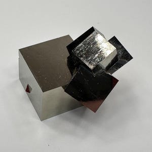 Könnte beinhalten: Ein natürliches Pyrit-Mineralexemplar mit mehreren miteinander verbundenen metallischen, messingfarbenen kubischen Kristallen. Die Ansammlung zeigt scharfe, geometrische Kanten und reflektierende, glatte Oberflächen mit einem ausgeprägten goldenen Glanz und einem kleinen roten dreieckigen Einschluss.