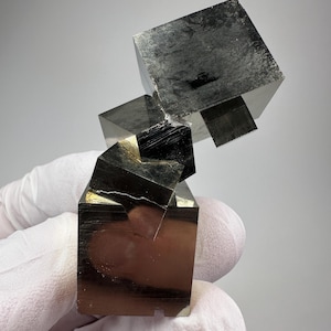 Könnte beinhalten: Ein Cluster aus Pyritwürfeln, einem metallischen Mineral, wird vor einem neutralen Hintergrund gezeigt. Die Würfel haben ein reflektierendes, messingartiges Aussehen mit scharfen Kanten und einer geometrischen Struktur. Die Probe wird von einer behandschuhten Hand gehalten.
