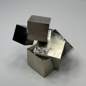Może przedstawiać: Skupisko metalicznych kryształów pirytu z wieloma połączonymi sześciennymi formacjami o odblaskowych, mosiężno-złotych powierzchniach i ostrych, geometrycznych krawędziach. Okaz minerału wykazuje naturalne, gładkie powierzchnie i wyraźne struktury.