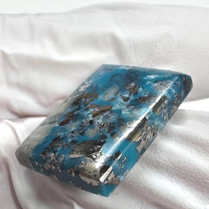 May include: A rectangular polished gemstone cabochon featuring a vibrant turquoise blue base colour with prominent metallic silver and dark grey inclusions distributed across the surface, showcasing a smooth, reflective, and glossy finish.