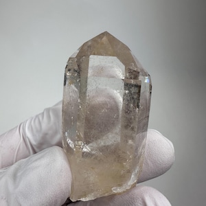 May include: A clear, hexagonal quartz crystal point with a pointed termination. The mineral specimen features natural internal inclusions, visible facets, and a translucent, pale brownish-yellow tint throughout its solid, geometric structure.