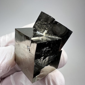 Könnte beinhalten: Ein polierter Pyritwürfel, auch bekannt als Narrengold, mit einem reflektierenden, metallischen Glanz. Der Würfel wird von einer behandschuhten Hand vor einem neutralen Hintergrund gehalten. Die geometrische Form des Minerals ist deutlich erkennbar.