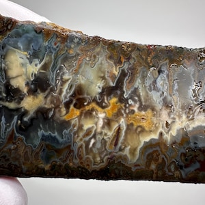 May include: A polished slab of agate stone featuring intricate, swirling patterns in shades of deep brown, charcoal gray, creamy white, and vibrant mustard yellow. The surface displays a complex, organic texture with translucent and opaque areas.