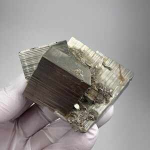 Op de afbeelding: Een metalliek pyriet mineraal exemplaar met duidelijke kubusvormige kristalformaties en een reflecterend, messinggoud oppervlak. Het cluster vertoont scherpe, geometrische randen en gestreepte texturen op de vlakken van de kubussen, wat een natuurlijke, ongepolijste uitstraling geeft met wat ruw, gemineraliseerd matrixmateriaal aan de zijkant.