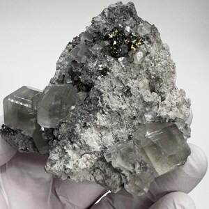 May include: A cluster of clear, green, and grey crystals with a metallic gold accent. The crystals are embedded in a grey, rocky matrix.