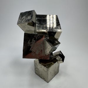 Op de afbeelding: Een metalen pyrietsculptuur bestaande uit gestapelde, onregelmatig gevormde kubussen. De sculptuur heeft een reflecterend, zilvergrijs oppervlak met zichtbare textuur en kleurvariaties. De opstelling creëert een dynamische, abstracte vorm.