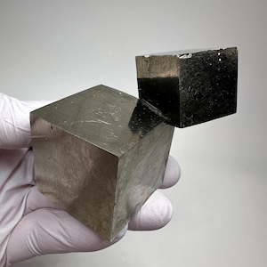 Op de afbeelding: Twee metalen pyrietkubussen, de ene groter dan de andere, worden getoond tegen een neutrale achtergrond. De kubussen hebben een reflecterend, gepolijst oppervlak, dat hun geometrische vorm laat zien. De grotere kubus is onder de kleinere geplaatst.