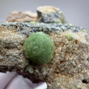 May include: Close-up of a mineral specimen featuring a vibrant green, spherical formation. The mineral is set against a backdrop of rough, textured rock with shades of brown, gray, and hints of green. The overall composition highlights the natural beauty of the mineral.