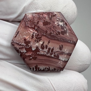 May include: A polished hexagonal gemstone cabochon featuring a smooth surface with layered bands of dusty rose, mauve, and deep burgundy colors. The stone displays intricate, dark dendritic fern-like inclusions scattered across the surface.