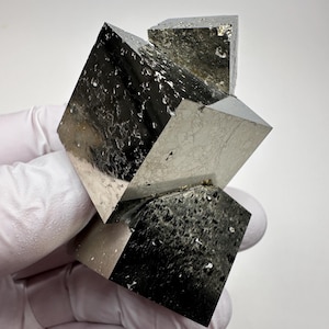 Könnte beinhalten: Ein Cluster aus Pyritwürfeln, einem metallischen Mineral, mit einem reflektierenden, silberartigen Aussehen. Die Würfel sind in einer visuell auffälligen Formation angeordnet, die ihre geometrische Präzision und natürliche Schönheit zeigt. Der Pyrit hat eine leicht strukturierte Oberfläche.