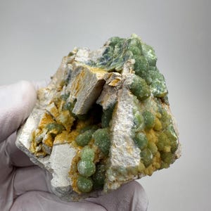 May include: A mineral specimen featuring clusters of small, rounded, translucent green crystals embedded within a rough, off-white matrix. The stone displays patches of yellow-orange mineral deposits nestled between the pale, angular rock formations.