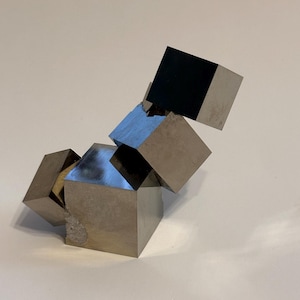 Könnte beinhalten: Eine Ansammlung natürlicher Pyritkristalle, bestehend aus mehreren metallischen, messingfarbenen Würfeln mit scharfen Kanten und reflektierenden Oberflächen. Die Mineralformation zeigt eine gestufte, geometrische Anordnung mit sichtbaren Kristallstrukturen.