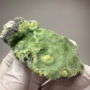 May include: A close-up of a vibrant green mineral specimen. The stone features a unique, layered pattern with circular formations in varying shades of green and yellow. The mineral has a rough, textured surface with dark gray and black accents.