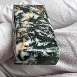 May include: A rectangular polished gemstone cabochon featuring a complex pattern of deep black, forest green, and creamy white mineral inclusions. The stone has a smooth, reflective surface with visible crystalline textures throughout its structure.