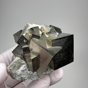 May include: A cluster of metallic pyrite crystals featuring sharp, cubic formations with a reflective, brassy gold surface. The mineral specimen is attached to a rough, light gray rocky matrix base, showcasing geometric shapes and distinct edges.