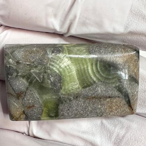 May include: A rectangular stone with a mix of gray, brown, and green hues. The stone has a polished surface with a swirling pattern of green and gray, and a rough, textured appearance.