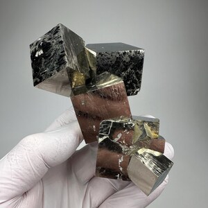 Op de afbeelding: Een cluster van pyrietkubussen, ook wel bekend als 'fool's gold', met een metallic glans. De kubussen zijn een mix van zilver, goud en zwart, met zichtbare kristallijne structuren. Het mineraal specimen wordt in een gehandschoende hand gehouden.