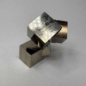 Op de afbeelding: Een cluster van metalen pyrietkristallen met meerdere onderling verbonden kubussen met een reflecterend, messinggoud oppervlak. Het mineraalexemplaar vertoont scherpe, geometrische randen en gladde, platte vlakken die het licht opvangen, waardoor er duidelijke highlights en schaduwen over de kubusvormige structuur ontstaan.