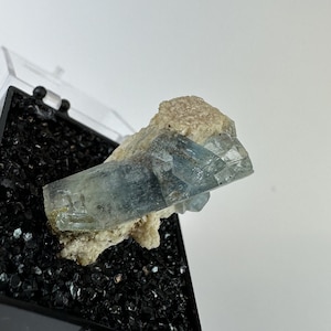 以下が含まれることがあります： 黒い砂利のベッドの上に置かれた青いアクアマリンの結晶標本。黒いディスプレイボックスに入っています。