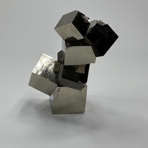 May include: A cluster of metallic pyrite crystals featuring multiple interconnected cubic formations. The mineral specimen displays a reflective, brassy gold surface with sharp, geometric edges and distinct natural facets throughout the structure.